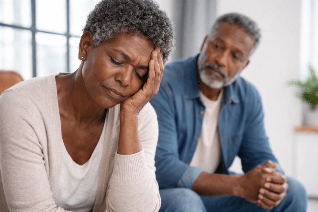 Elderly African American woman sitting with her head in her hand looking distressed due to menopause misdiagnosis, while her husband sits nearby watching her with concern